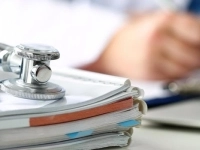 Stéthoscope posé sur une pile de dossiers médicaux, avec une personne écrivant à l’arrière‑plan.