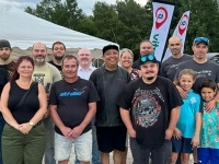 Groupe de personnes réunies à l’extérieur devant un chapiteau blanc, avec des bannières et une zone boisée en arrière-plan.