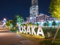 Vue nocturne d’un espace urbain à Osaka, avec de grandes lettres illuminées formant le mot « OSAKA » et des immeubles éclairés en arrière‑plan.