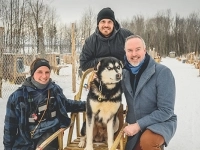 Trois personnes posant à l’extérieur dans un décor enneigé avec un grand chien de traîneau assis sur un traîneau, devant un chenil hivernal.
