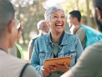 Personne tenant une tablette numérique et discutant avec un petit groupe en plein air, dans un environnement boisé et ensoleillé.