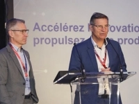 Deux personnes en tenue professionnelle se tenant sur une scène, l’une derrière un lutrin, devant un écran affichant un message sur l’innovation et la productivité. 