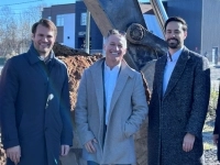 Groupe de personnes debout sur un chantier devant une pelle mécanique et un tas de terre, avec des bâtiments industriels en arrière-plan.