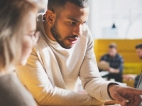 Personne pointant un écran d’ordinateur dans un environnement de bureau moderne, avec d’autres personnes en arrière-plan.
