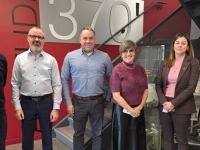 Groupe de personnes posant dans un espace industriel moderne, devant un mur rouge portant l’inscription « 370! », près d’un escalier métallique et de vitrines présentant des pièces mécaniques.
