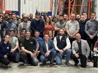 Grand groupe de personnes posant pour une photo à l’intérieur d’un entrepôt industriel, entouré de rayonnages métalliques orange et d’équipements de manutention.